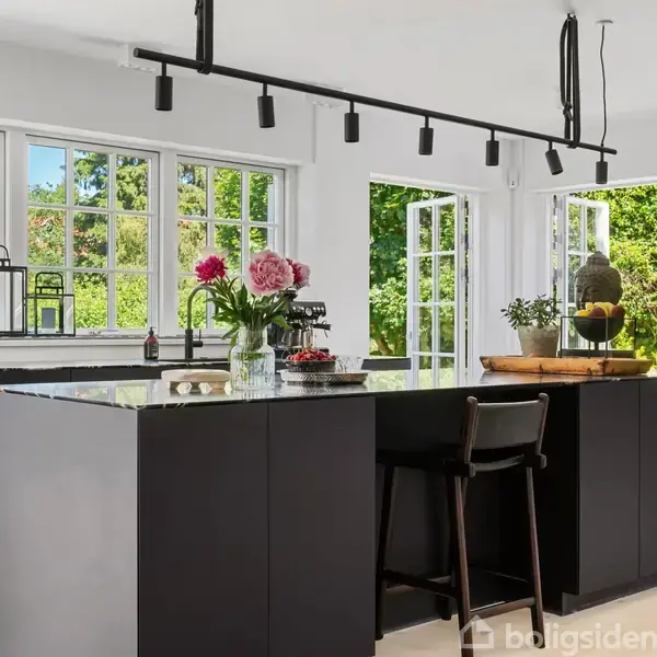 Et moderne, sort køkkenbord med blomster og frugter står i et lyst køkken. Vinduerne åbner ud til en grøn have. En høj, sort skænk er placeret i baggrunden.