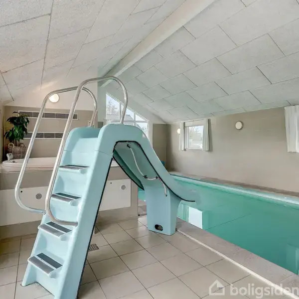 En indendørs swimmingpool med en lyseblå rutsjebane, omkranset af flisegulv og lyse vægge, oplyst af naturligt lys fra vinduer. Der er ingen tekst i billedet.