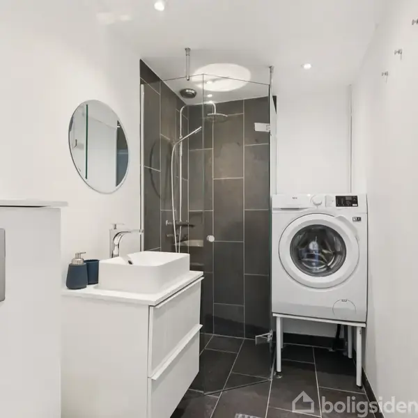 Vaskemaskine står i moderne badeværelse med brusekabine og håndvask. Rummet er lyst med sorte gulvfliser, rundt spejl over vasken og vægmonteret toiletknap.