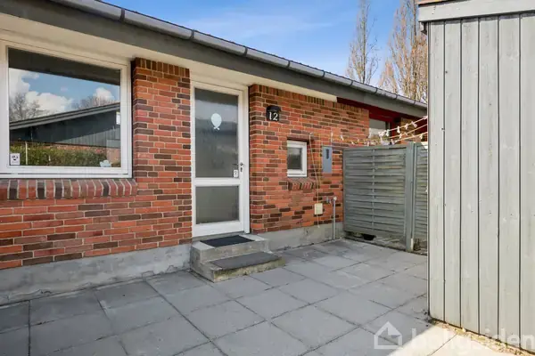 Murstenshus med hvid hoveddør og trappe foran. Husnummer 12 ved siden af døren. Omkringliggende flisebelagt gårdhave og træhegn mod højre, klar blå himmel i baggrunden.