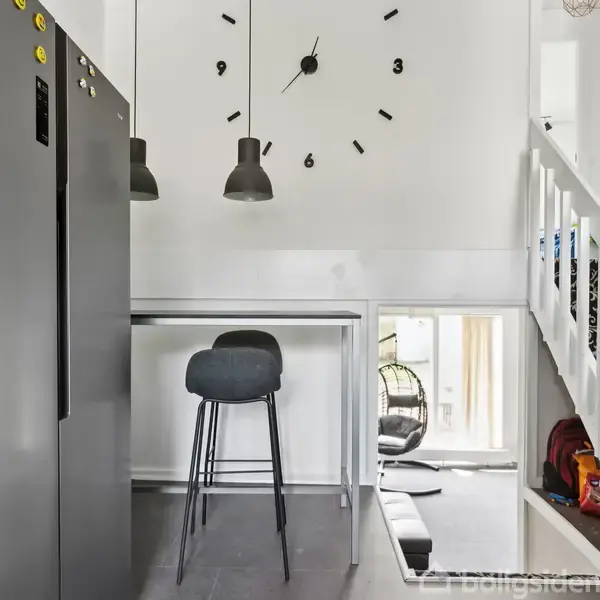 Et stort ur på en hvid væg viser klokken 3. Et sort skab står ved siden af en barstol ved et højt bord. Trappe og lamper er i nærheden.