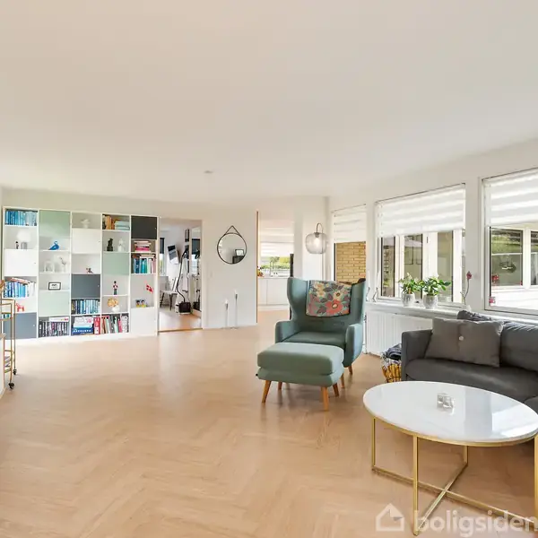Lyst stueområde med grå sofa og grøn lænestol, placeret omkring et rundt sofabord. Store vinduer bringer lys ind, og indbygget hylde fylder bagvæggen med bøger og dekorative objekter.