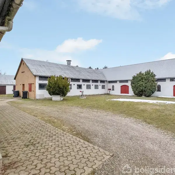 En gårdsplads med tre store staldbygninger i hvid og beige, omgivet af grønne buske. Tætsluttende tagkonstruktioner ses under en klar blå himmel med enkelte skyer.