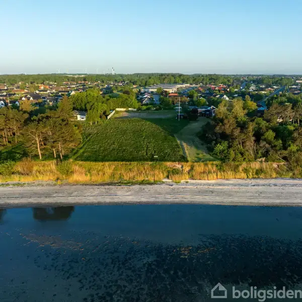 Strand grænser op til græsområde med træer, bagved ligger en by med huse blandt grønne landskaber og blå himmel i baggrunden.