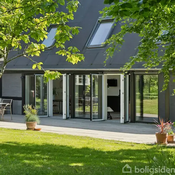 Huset har åbne dobbeltdøre ud mod en terrasse med havemøbler. I forgrunden er der grønne græsplæner og træer, der skaber en skyggefuld atmosfære.