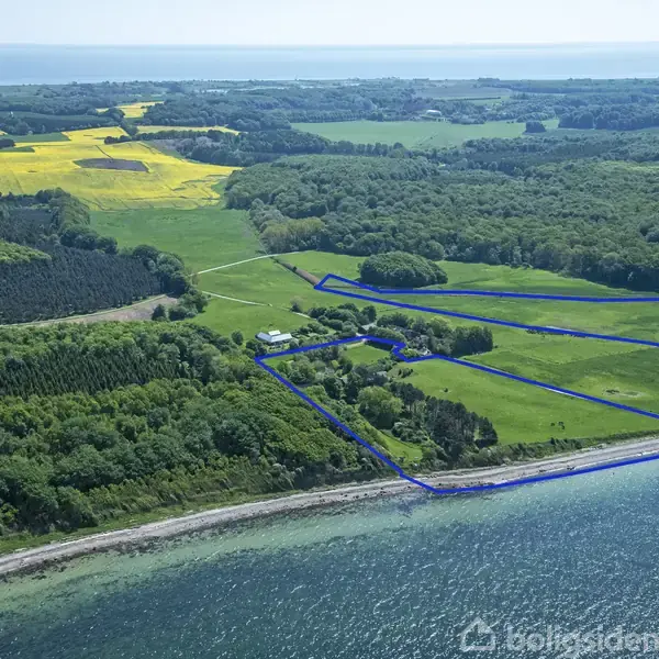 Et landområde med marker omgivet af skov og hav. Blå linjer markerer grænserne. Landskabet inkluderer marker, skovområder og kystlinje langs vandet. Ingen tekst bortset fra "Boligsiden".