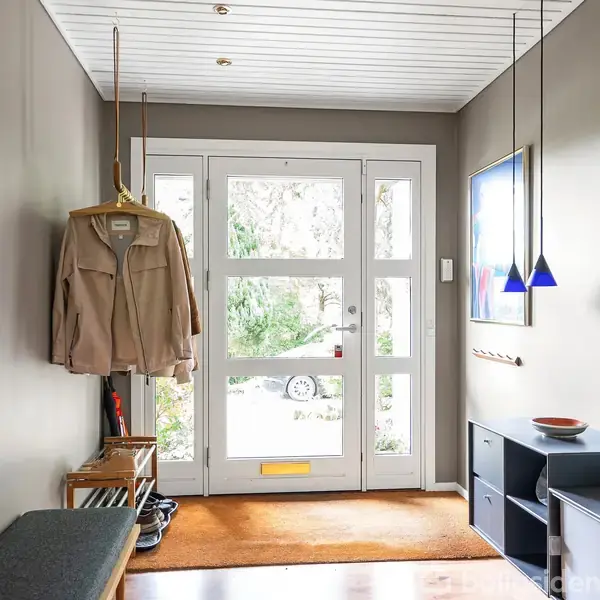 Frakke hænger på bøjle i entré. Skænke og belysning supplerer rummet med store glasdøre, spejl på væggen og planter, der skaber en indbydende atmosfære.