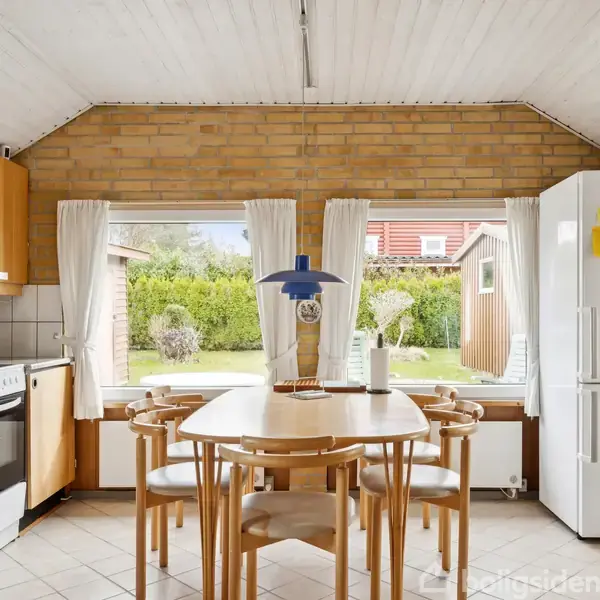 Et enfamiliehus med gul murstensfacade har en terrasse foran, hvor der står grønne havestole og borde. Haven er omgivet af hække, og træer ses i baggrunden.