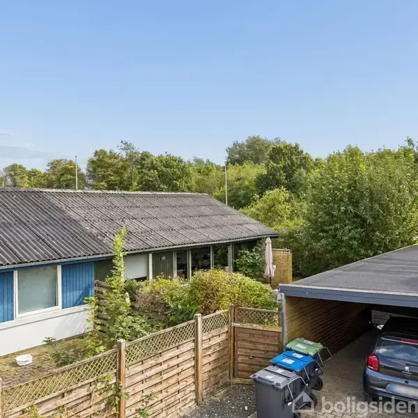 En fladt tagbygning ved siden af en carport, der rummer en bil. Omkring er der træer og en hæk ved hegnet samt skraldespande foran.