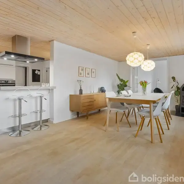 Spisebord med stole står i en moderne stue. Rummet har et åbent køkken i baggrunden, en brændeovn i hjørnet og lyse trægulve. To pendellamper hænger over bordet.