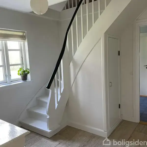 En hvid trappe med sort gelænder fører opad i en lys entré. En potteplante står på en vindueskarm ved siden af trappen. Døren til højre er lukket.