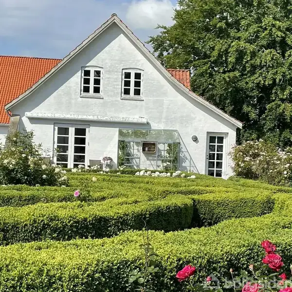 Hvidt hus med rødt tegltag står bag veltrimmet hæk i snoede mønstre. Glasudestue til venstre. Omgivet af blomster og træer under en klar blå himmel.