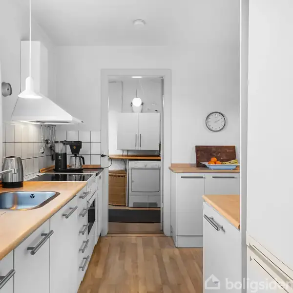Et lyst køkken med hvide skabe og træbordplader. Køkkenet har en vask, kogeplade og kaffemaskine. En vægklokke hænger, og et skærebræt med frugt ses i baggrunden.