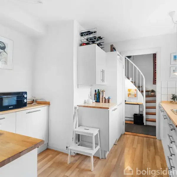 Køkken med hvide skabe og træbordplader, hvor en mikrobølgeovn står. En lille stige er placeret ved siden af skabene. En trappe fører til et andet rum.