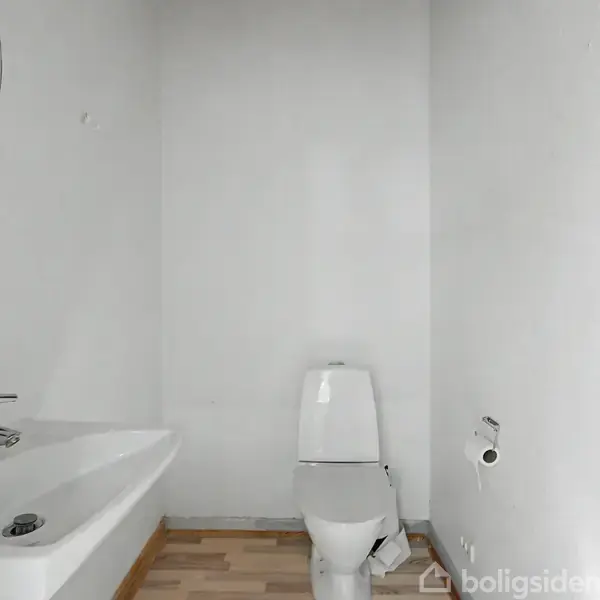 Toilet og håndvask er placeret i et hvidt, lille badeværelse. Toiletpapirholderen er tom, og væggene er uden dekorationer, hvilket skaber et enkelt udseende. Gulvet har lyst trægulv.