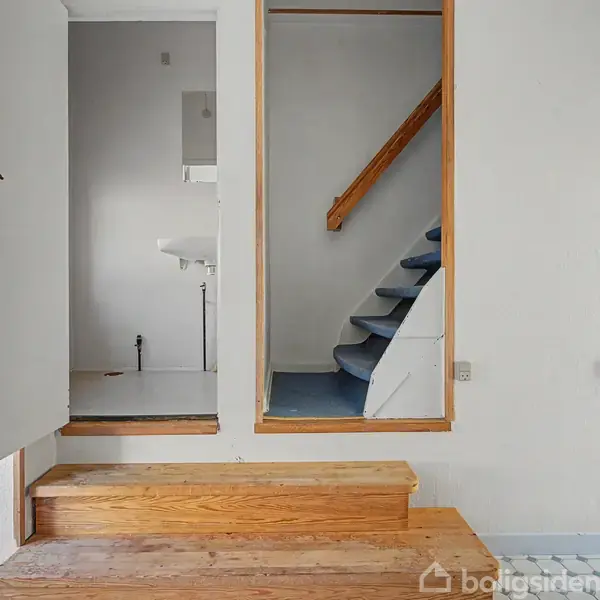 Dør står åben til rum med håndvask, trapper fører opad, omgivet af lyse vægge og gulv. Radiator placeret til højre.