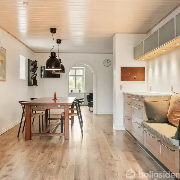 Køkken med træbord og stole under sorte lamper; til højre er der moderne køkkenmoduler med indbygget belysning; træ- og hvidmalet indretning skaber en lys atmosfære.