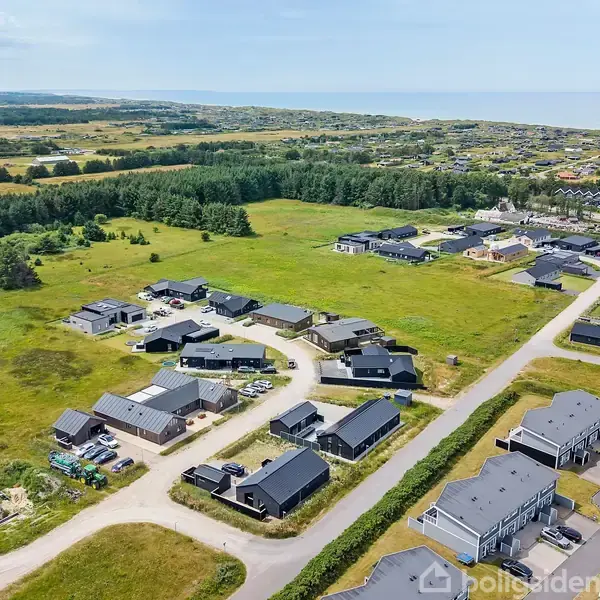 Bebyggelse med moderne huse ligger spredt i en forstad. Husene er omgivet af åbne grønne marker, og i det fjerne ses en skov og flere tætte boligområder.
