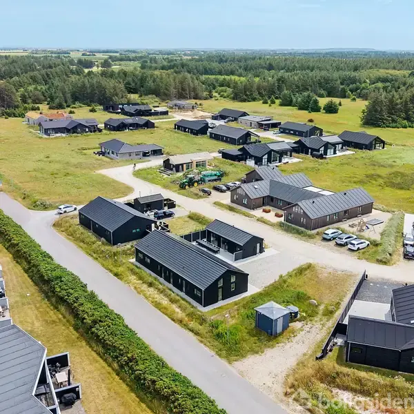 Boliger med sorte tage ligger spredt i et grønt boligområde med trær i baggrunden, omgivet af veje og græsarealer under en klar blå himmel.