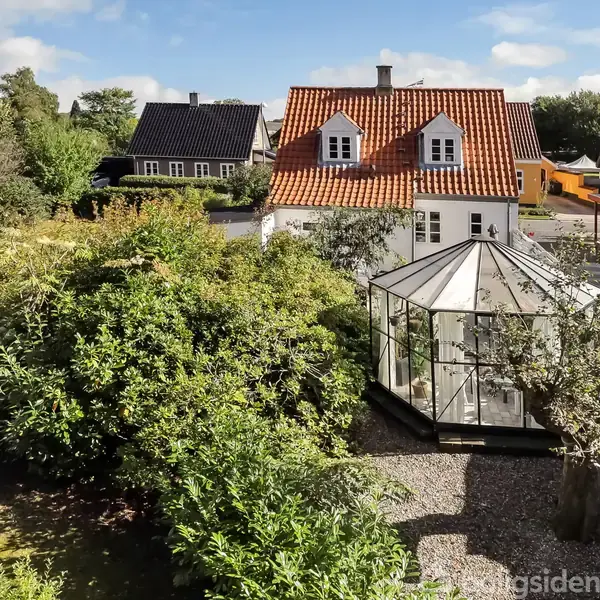 Et hvidt hus med rødt tag står i en frodig have med en drivhusstruktur. Omgivelserne inkluderer grønne planter, træer og nabohuse i et landsbyagtigt miljø.