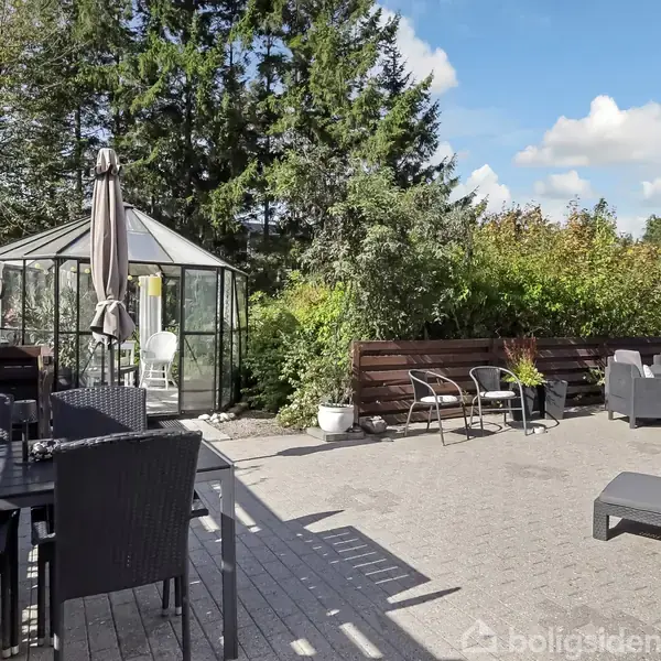 Et udendørs terrasseområde med flisebelagt gulv har sorte havemøbler, liggestole og et drivhus. Omgivet af grønne planter og træer, under en blå himmel med skyer.
