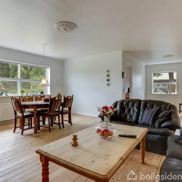 Stue med træbord og sofa i forgrunden, spisested med bord og stole i baggrunden. Lyse vægge og trægulv giver rummet en hyggelig atmosfære.