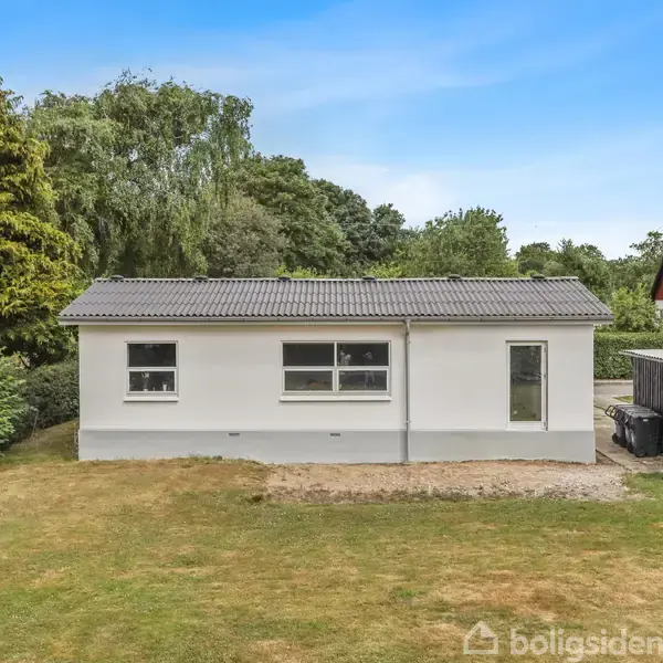 Et lille, hvidt hus med gråt tag står i en grøn have med hække og træer omkring. En grussti løber langs siden, og der er en carport til højre.