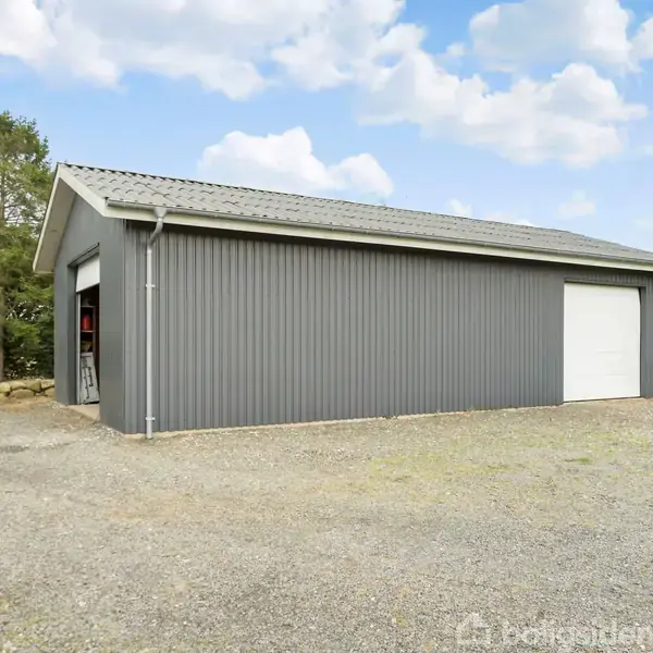En grå metalbygning står stille på en grusbelagt plads. I baggrunden ses træer og en blå himmel med skyer. En lukket port og en åben dør er synlige.