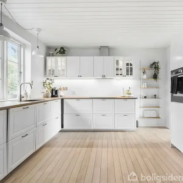 Et moderne køkken med hvide skabe og træbordplader. Stort vinduesparti til venstre slipper naturligt lys ind. Indbygnings-ovne er integreret i et mørkt skab til højre.