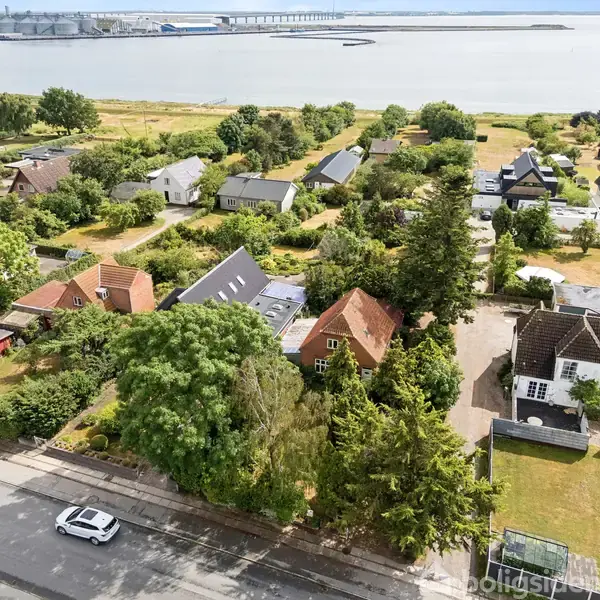 Huse ligger blandt træer og buske i en grøn bydel med udsigt over vandet. En vej løber foran med en hvid bil parkeret.