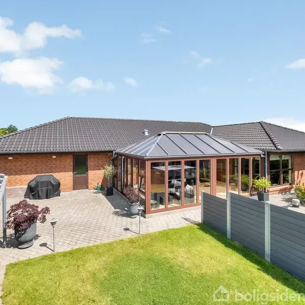 Rødt murstenshus med sort tag har en stor terrasse med grill og havemøbler. En glasveranda er centralt placeret, omgivet af grøn have og grå hegn. Solrig, blå himmel.