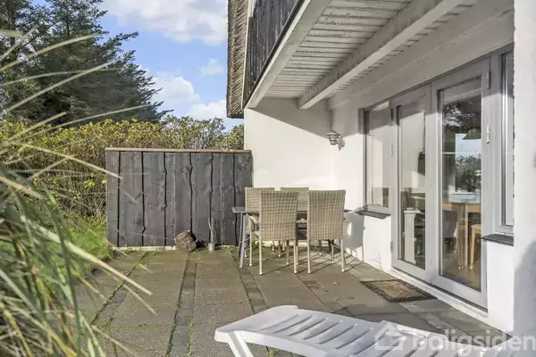 En terrasse med et bord og fire stole står i skyggen af et hus med store vinduer. Omkring terrassen er der planter og en blå himmel kan ses i baggrunden.