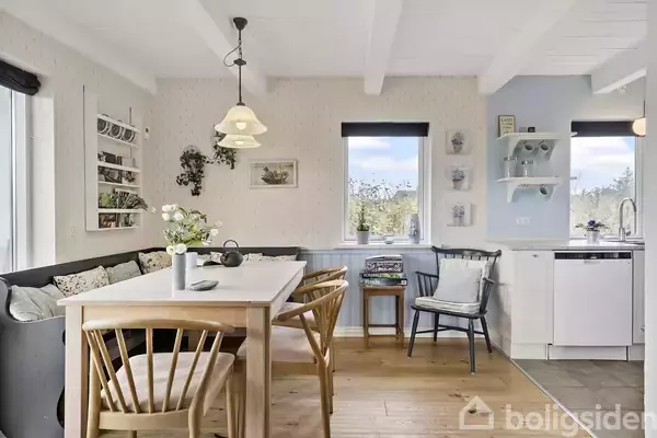 Spisebord med træstole står i et lyst køkkenalrum. Lampe hænger over bordet, omgivet af planter og billede på væggen. Køkken med opvaskemaskine ses i hjørnet.