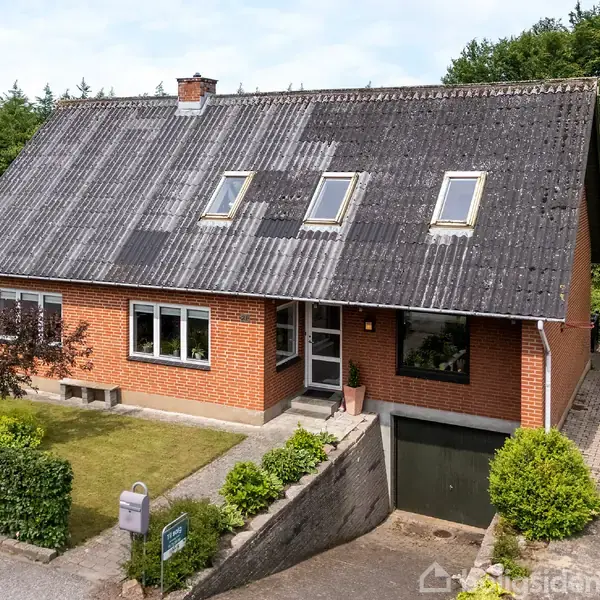 Rødt murstenshus med gråt skråt tag og flere tagvinduer. Foran huset er en velholdt have med træer og buske. Garagen er integreret i kælderniveauet.