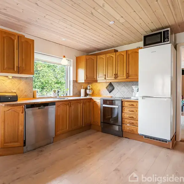 Køkken med træskabe indeholder en ovn, opvaskemaskine og køleskab. En mikroovn står ovenpå køleskabet. Knive hænger på væggen, og et stort vindue giver udsigt til grønne træer.