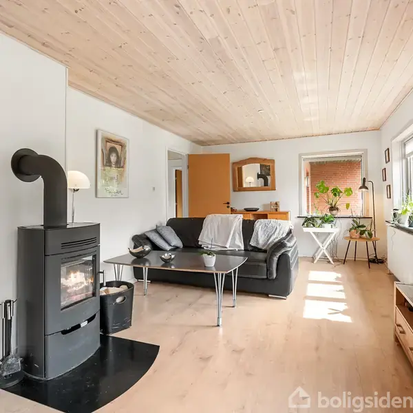 Brændeovn opvarmer stue med grå sofa, sofabord og moderne indretning. Lysindfald fra vinduer skaber hyggelig atmosfære. Grønt område ses gennem åbent dør, billeder hænger på væggen.