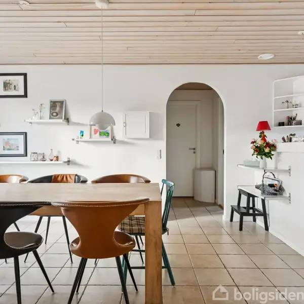 Et spisebord med forskellige farvede stole står i en lys spisestue. Der er hylder med dekorationer på væggen, hvide skabe og en åbning til et tilstødende rum.