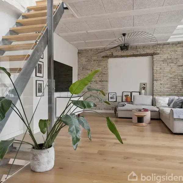 En stor grøn plante står ved siden af en moderne trappe med glasgelænder. I baggrunden en stue med lys sofa, vægdekorationer og murstensvægge, der skaber et lyst og stilrent miljø.