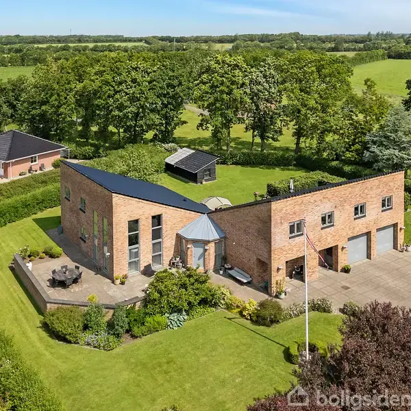 Industrielt designet to-etagers murstenshus med fladt tag og store vinduer, omgivet af en velplejet have med terrasse og omkranset af høje træer på en stor græsplæne i landlige omgivelser.