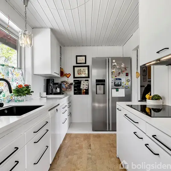 Et moderne køkken med hvide skabe er udstyret med en sort vask og kogeplade. Et køleskab står for enden, omgivet af lyst træ og et vindue med blomstret gardin.