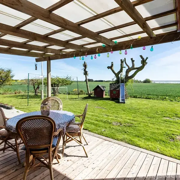 Terrassemøbler står på en overdækket træterrasse. Farverige lamper hænger fra loftet. I baggrunden er en stor græsplæne med klatrestativ, et lille hus og åbne marker.