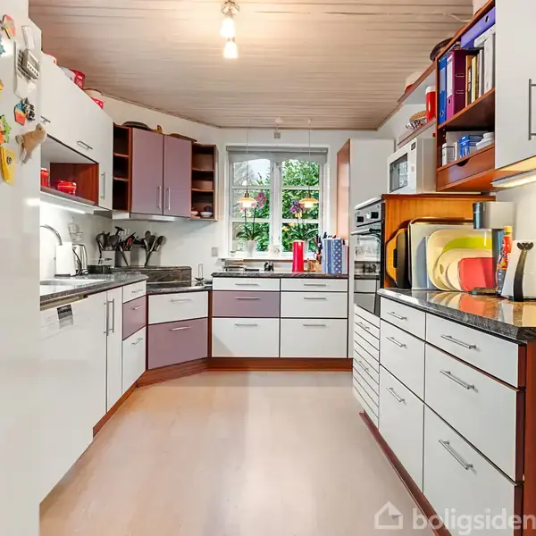 Køkken med hvide skabe, trædetaljer og sorte bordplader, fyldt med køkkenudstyr. Et stort vindue lader lys ind, omgivet af hylder med bøger og planter. Køleskab har magneter.