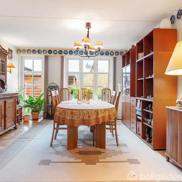 Et spisebord dækket med dug står midt i et lyst spisestue oplyst af en lysekrone; omgivet af træmøbler, grønne planter ved vinduer og en stor lampe på en skænk.