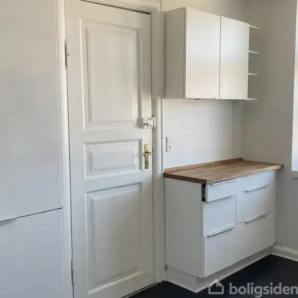 Hvidt køkken med skabe og en bordplade ved siden af en dør. En stor vindue lader naturligt lys ind i rummet.