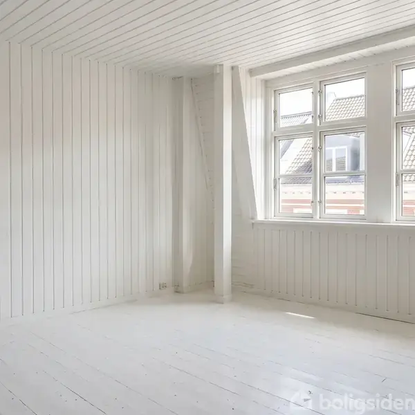 Et lyst rum har hvide træpaneler og er tomt, med store vinduer mod højre, som giver udsigt til hustage udenfor.