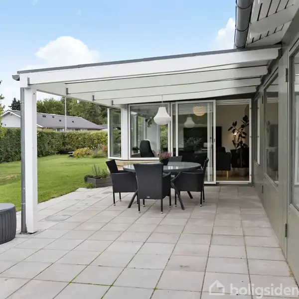 Overdækket terrasse med spisebord og stole står på flisebelægning, omgivet af velplejet have med græsplæne og buske. Der er en sort puf ved siden af.