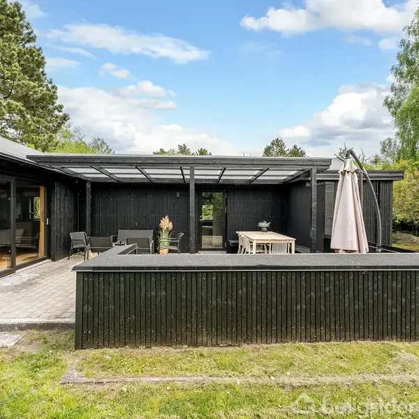Sort træhus med stor terrasse, møbleret med borde og stole under en pergola. Omgivet af grønne træer og græsplæne under blå himmel med skyer.
