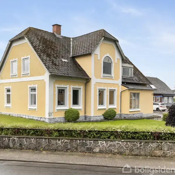 Et gult hus med hvide detaljer står roligt. Det har en forhave med trimmet græs og buske, omgivet af en lav stenmuret kant. Himlen er blå med få skyer.