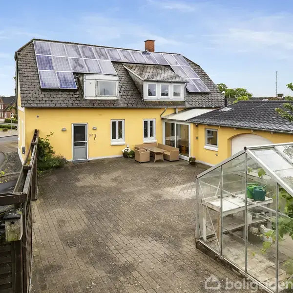 Gult hus med solpaneler på taget, står ved en brolagt gårdsplads. Til højre ses et drivhus. Nabohuse og grønne buske omkranser gårdspladsen.