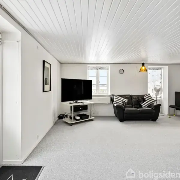 Et lyst stueområde med en sort lædersofa ved et vindue. Til venstre står et tv på et stativ. Reol og stol ses i baggrunden.